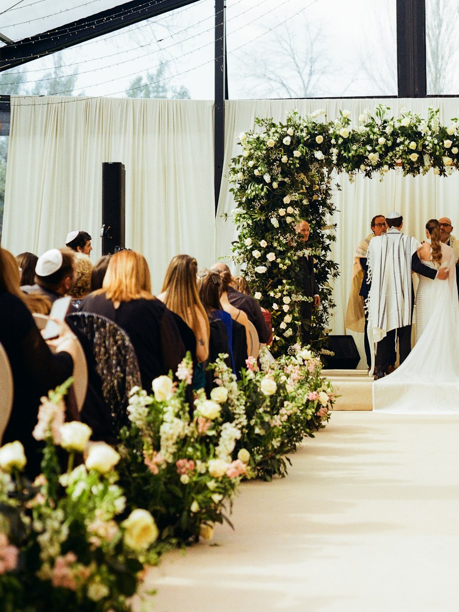 A couple under a floral chuppah at a wedding ceremony, guests seated along a flower-lined aisle